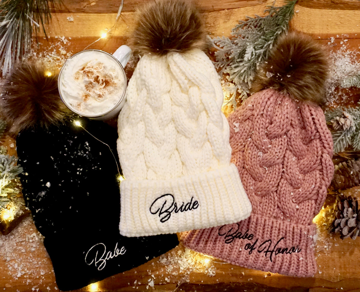 Three bachelorette beanies displayed on a piece of natural wood with fake snow, twinkle lights, and a cup of hot chocolate. The first beanie is ivory with "bride" embroidered in black, the second beanie is mauve with "babe of honor" embroidered in black, and the third beanie is black with "babe" embroidered in white.