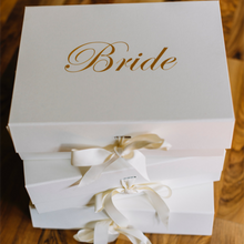 Load image into Gallery viewer, An ivory Personalized Bride Gift Box with a closed lid, featuring the word "Bride" in a gold chrome script font. The box is finished with a grosgrain ribbon and has a magnetic closure. The empty interior of the box is ready to be filled with goodies, and the lid can be customized with a message option. The elegant and simple design of the box makes it a timeless keepsake for any Bride-to-be.