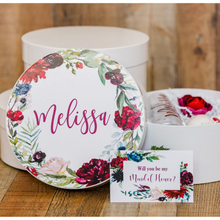 Load image into Gallery viewer, A white round gift box with a lid, featuring a dark floral print and personalized name decal adhered to the top of the box. The box is shown with a white crinkle paper filling and a ribbon tied around it.