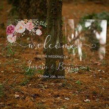 Load image into Gallery viewer, A breathtaking clear acrylic wedding sign adorned with beautiful florals is set against a lush greenery backdrop, creating a picturesque display. The sign reads &quot;Welcome to our wedding,&quot; written in a romantic script font, with the names of the couple featured prominently below.
