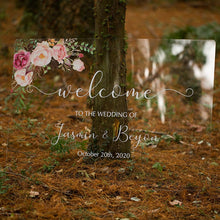 Load image into Gallery viewer, A breathtaking clear acrylic wedding sign adorned with beautiful florals is set against a lush greenery backdrop, creating a picturesque display. The sign reads "Welcome to our wedding," written in a romantic script font, with the names of the couple featured prominently below.