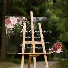 Load image into Gallery viewer, Against a backdrop of lush greenery, a clear acrylic wedding sign decorated with delicate florals welcomes guests to the celebration. The sign reads "Welcome to our wedding," written in an elegant script font, with the names of the happy couple featured prominently below.
