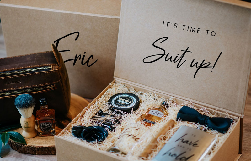 Kraft gift box with the name on top and the phrase "it's time to suit up" on the inside lid. The lid is open to reveal small gifts nestled in natural filler.