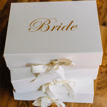 Load image into Gallery viewer, An ivory Personalized Bride Gift Box with a closed lid, featuring the word &quot;Bride&quot; in a gold chrome script font. The box is finished with a grosgrain ribbon and has a magnetic closure. The empty interior of the box is ready to be filled with goodies, and the lid can be customized with a message option. The elegant and simple design of the box makes it a timeless keepsake for any Bride-to-be.

