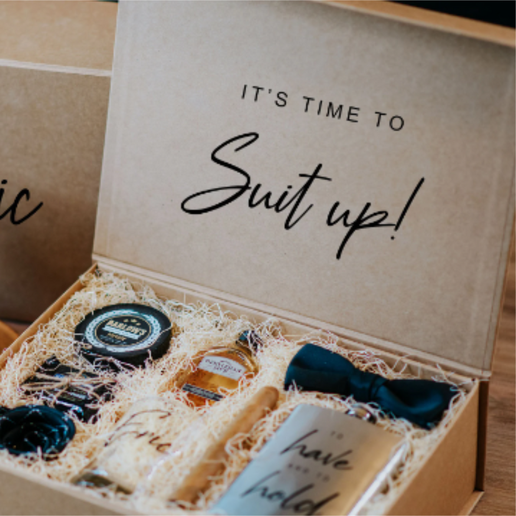 An image of a groomsmen gift box, crafted from kraft material with the phrase 'It's time to suit up' on the lid. The box is filled with natural filler and contains a variety of gifts including cufflinks, a pocket square, a tie clip, and a flask. These gifts are carefully arranged inside the box, creating a perfect groomsmen gift box presentation.