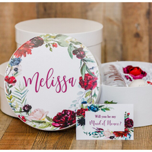 Load image into Gallery viewer, A white round gift box with a lid, featuring a dark floral print and personalized name decal adhered to the top of the box. The box is shown with a white crinkle paper filling and a ribbon tied around it.
