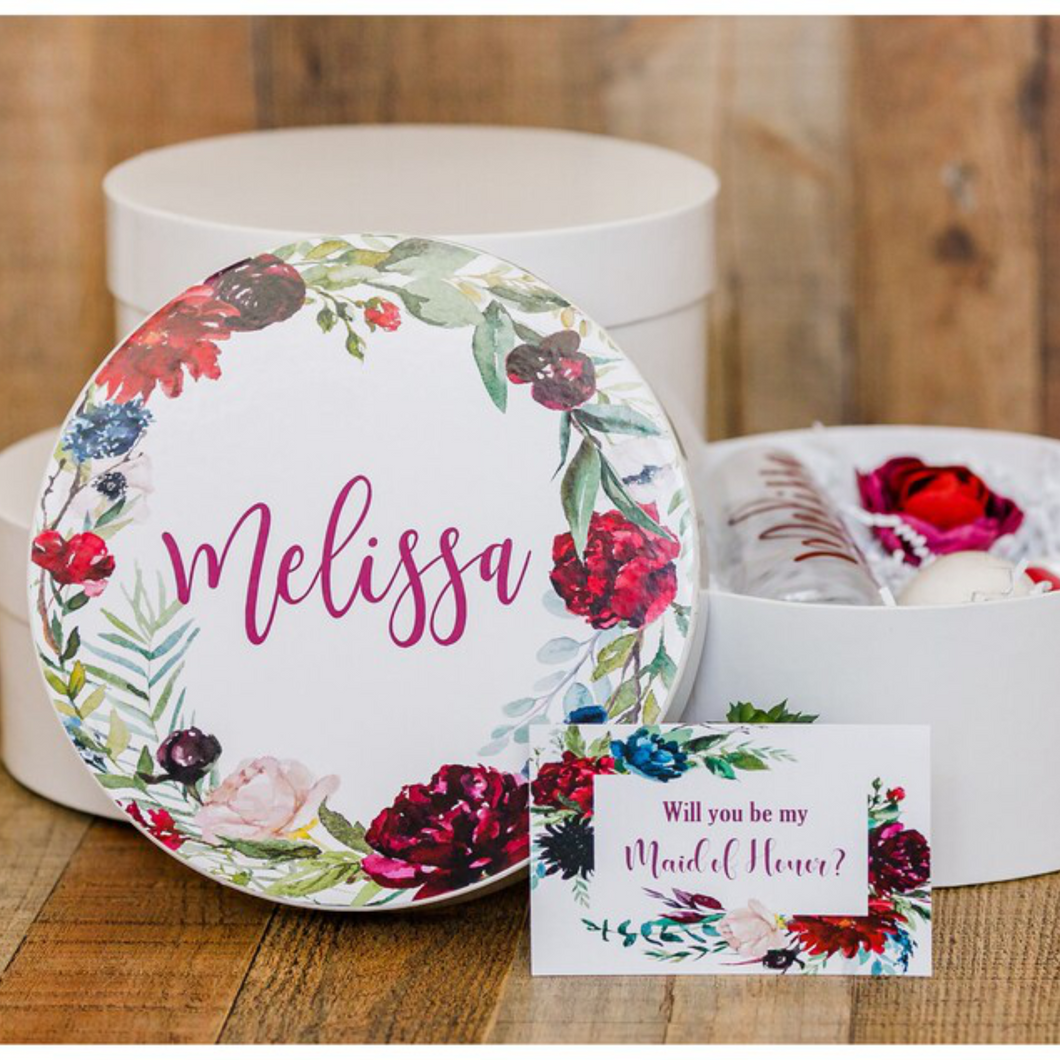 A white round gift box with a lid, featuring a dark floral print and personalized name decal adhered to the top of the box. The box is shown with a white crinkle paper filling and a ribbon tied around it.