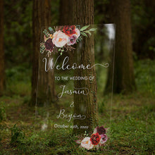 Load image into Gallery viewer, A gorgeous clear acrylic wedding sign surrounded by vibrant greenery and beautiful florals invites guests to celebrate the special day. The sign reads &quot;Welcome to our wedding,&quot; written in a stunning script font, with the names of the couple featured prominently below.
