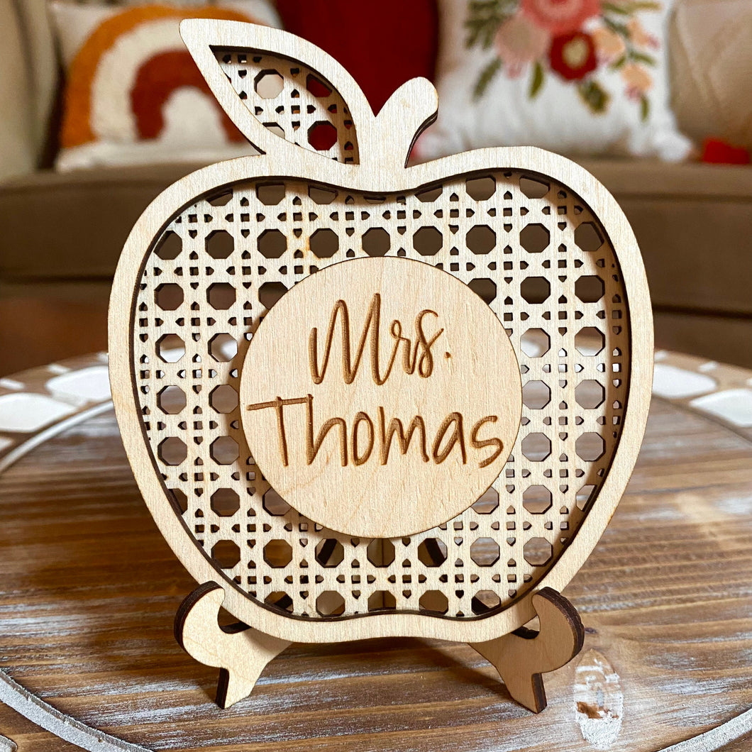 An intricately designed, laser-cut cane apple with a self-locking stand, perfect for use as a teacher name plate. The design is boho-inspired and features an engraved name in the center. This personalized decor piece can be placed on a desk or bookshelf, and makes for an adorable and practical teacher gift.